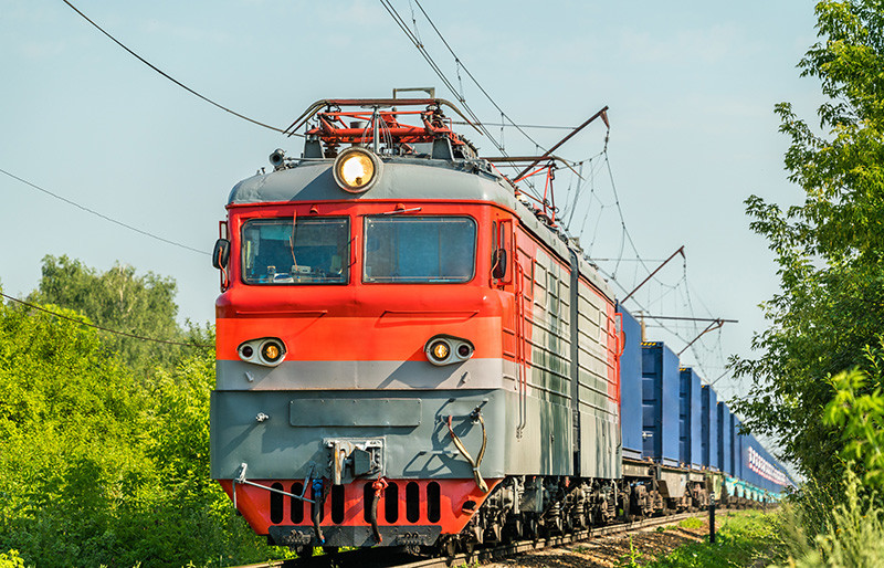 Železniční doprava - přední pohled na elektrickou lokomotivu a kontejnerový vlak projíždějící zelenou krajinou